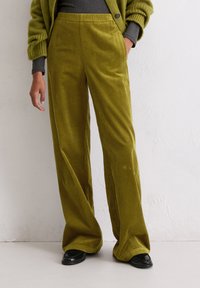 Groene corduroy wijde broek met verticale ribbels en een elastische tailleband. Gekleed met een donkergrijze top en zwarte schoenen.