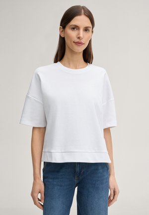 Kvinde med glat brunt hår iført en simpel hvid kortærmet skjorte og blå jeans, stående foran en lysegrå baggrund.