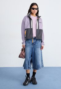 Purple and gray fleece jacket with zipper, pink t-shirt, wide denim culottes, black sneakers, and a small brown handbag.