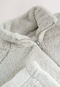 Veste polaire douce gris clair avec col haut et fermeture éclair argentée partiellement ouverte, montrant les étiquettes du vêtement à l'intérieur.