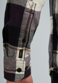 Karierte Flanelleute in Dunkelpurpur, Creme und Braun. Verfügt über schwarze Knöpfe, ein Markenetikett und einen strukturierten Stoff.