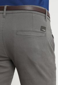 Grå byxor med en slät textur, med en bakficka och ett brunt läderbälte. Tyget är hållbart med en avslappnad design.