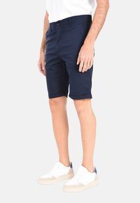 Shorts in cotone blu navy con taglio sartoriale, dotati di chiusura anteriore con bottone e orli arrotolati, abbinati a sneakers bianche e beige.