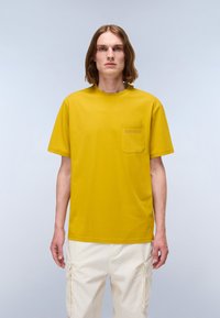 Person with shoulder-length brown hair wearing a mustard yellow T-shirt with a chest pocket and white cargo pants standing against a plain background.