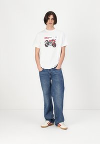 Hvid grafisk t-shirt med motorcykeltryk, parret med løse blå jeans og cremefarvede sneakers med røde striber. Enkel, afslappet design.