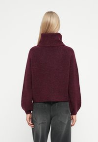 Maglione bordeaux con un col alto a coste, spalle scese e un design cropped. Materiale a maglia morbido e testurizzato abbinato a pantaloni grigio scuro.