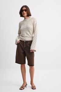 Cream striped knit sweater with long sleeves, paired with brown denim shorts and strappy black sandals, standing against a plain background.