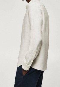 Vaaleanbeige pellavapaita, jossa on napitettava kaulus, pitkät hihat ja rento istuvuus. Huomattava rakenne ja sileä pinta. Yhdistetty tummiin housuihin.