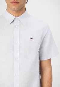 Kortsleeves skjorte med knapper i lyseblå og hvite striper, med en liten brodert logo i rødt, hvitt og blått på brystet.