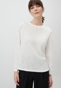 Witte lange mouwen top met een textuur van bloemmotieven, ronde halslijn en een subtiele glans, gecombineerd met zwarte broeken.