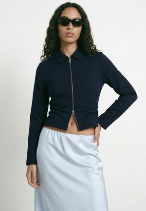 Femme aux longs cheveux bouclés portant des lunettes de soleil noires en forme d’œil de chat, une veste zippée bleu marine et une jupe en satin bleu clair, debout devant un fond uni.
