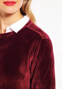 Top de terciopelo burdeos con cuello redondo, que presenta una camisa blanca con cuello debajo. Textura suave, mangas largas y detalles de hombros sin costuras.