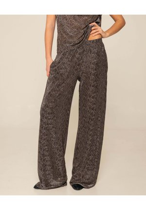 Mujer con pantalones anchos de pierna ancha marrones texturizados, con la mano en el bolsillo y parte superior a juego, de pie contra un fondo beige liso.