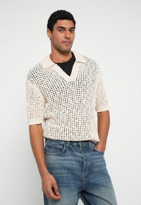 Suéter de manga corta tejido en color crema con patrón de malla negra, que presenta un escote en V y un cuello en contraste. Combinado con jeans de mezclilla azul.