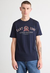 Tricou din bumbac de culoare bleumarin, cu guler rotund, având un imprimeu grafic cu "GANT 1949" și accente cu logo în roșu și alb. Croială dreaptă.