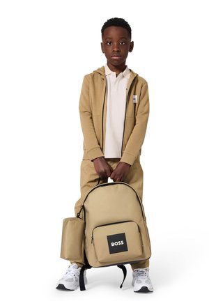 Beige rugzak met logo, ritssluiting en zwarte accenten. Gebruiker draagt een beige trainingspak en witte sportschoenen.