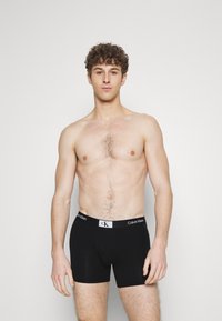 Juodos medvilninės boxer kelnaitės su baltu Calvin Klein logotipu ant juosmens. Lygus paviršius, prigludusi forma, iki šlaunų vidurio ilgio. Nėra jokių papildomų raštų.