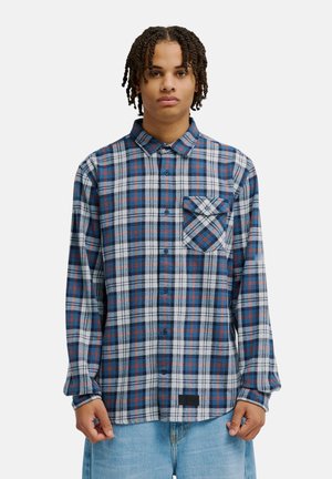 Jeune homme aux cheveux mi-longs torsadés portant une chemise à carreaux bleue, blanche et rouge à manches longues boutonnée et un jean bleu clair.