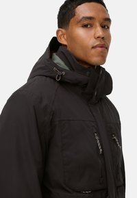 Schwarze isolierte Jacke mit hohem Kragen und Kapuze. Verfügt über Reißverschlusstaschen und kontrastierendes helles Futter. Strukturiertes Materialfinish.