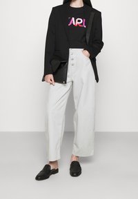 Blazer negro sobre una camiseta negra con un logo rosa, pantalones de pierna ancha gris claro con detalle de botones y zapatos negros de tipo slip-on.