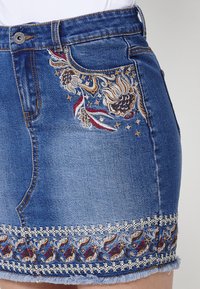 Jupe en denim ornée de broderies florales en or, rouge et bleu. Ourlet effiloché avec des détails en dentelle raffinés le long du bord inférieur.
