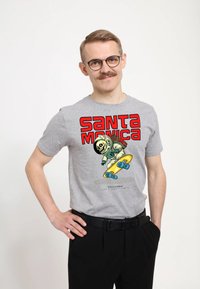 Muž s brýlemi a knírkem se usmívá, má na sobě šedé tričko s potiskem lebky astronauta na skateboardu a červeným textem "Santa Monica".