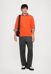 Lys orange tekstureret sweater, lagd over en lys lilla skjorte, parret med grå plisserede bukser og sorte sko med hvide accenter.