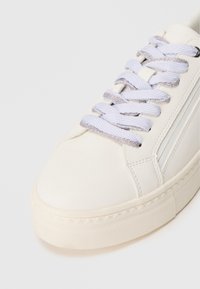 Λευκό δερμάτινο αθλητικό παπούτσι με αντίθετους ανοιχτό μπλε και μεταλλικού ς ασημί κορδόνια, διαθέτοντας υφή από καουτσούκ στη σόλα και μινιμαλιστικά σχεδιαστικά στοιχεία.