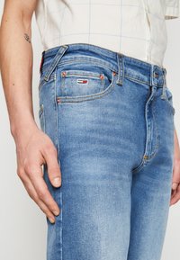 Vaqueros de mezclilla azul claro con un diseño clásico, costuras visibles, bolsillos frontales y un pequeño detalle de logo en la cinturilla.