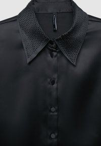 Schwarzes Satinhemd mit einem mit Nieten besetzten Kragen, sechs Knöpfen vorne und einer glatten Textur. Der Kragen ist mit kleinen, glänzenden Akzenten verziert.
