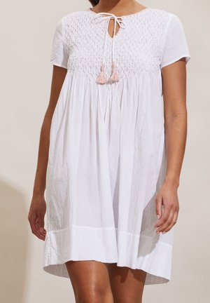 Femme portant une robe blanche à manches courtes avec un corsage texturé et un lien à pompons roses au niveau de l'encolure, se tenant devant un fond uni.