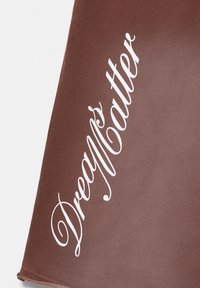 Tissu marron avec texte blanc en italique disant « Dreams Matter » en diagonale sur la surface.