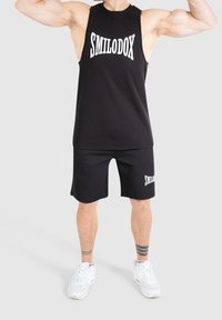 Débardeur noir sans manches et short assorti, tous deux avec le logo "SMILODOX" en blanc. Le tissu semble lisse ; coupe athlétique avec une grande liberté de mouvement pour les bras.