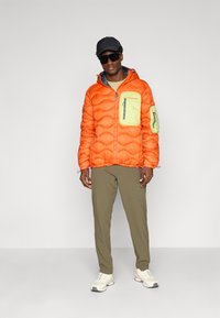 Oranje pufferjack met een groene borstzak, verticale rits en een gestructureerd, gewatteerd ontwerp. Gecombineerd met olijfgroene broek en sneakers.