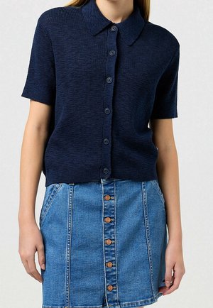 Kobieta ubrana w granatową krótka koszulkę na guziki z krótkim rękawem oraz niebieską spódnicę dżinsową z kieszeniami, stojąca na tle jednolitego tła.