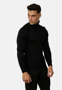 Un pull col roulé noir au design ajusté, fabriqué dans un matériau tricoté lisse. Manches longues et poignets côtelés, porté avec un pantalon sombre.