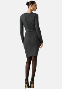 Abito aderente in maglia grigio scuro con maniche lunghe, dettagli a coste e vita increspata. Indossato con collant neri e tacchi.