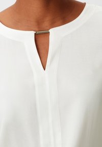 Blouse blanche avec un col en trou de serrure orné d'une barre métallique. Confectionnée en tissu léger avec une texture lisse et un éclat subtil.