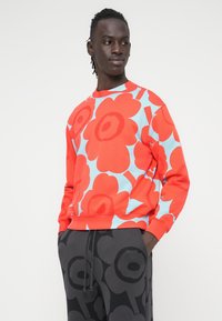 Κόκκινο φούτερ με λουλουδάτο μοτίβο και μπλε λεπτομέρειες, φτιαγμένο από μαλακό ύφασμα. Συνδυάζεται με μαύρο παντελόνι με παρόμοιο λουλουδάτο σχέδιο.