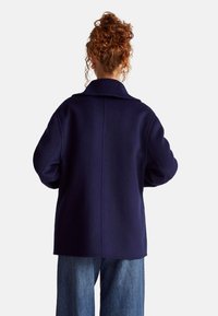 Cappotto in misto lana blu navy con collo a risvolto, spalle scese e vestibilità rilassata. Presenta una cucitura centrale ed è abbinato a denim a gamba larga.