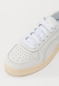 Baskets en cuir blanc avec embout perforé, lacets plats blancs et semelle en caoutchouc beige. Présente des panneaux texturés et un logo sur la languette.