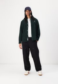 Cămașă verde din catifea cu milanezi peste un tricou alb, asociată cu pantaloni negri largi și adidași albi. O șapcă bleumarin completează ținuta.