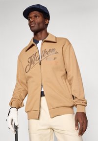 Brūna zip-up jaka ar ribotu apkakli, ar "Malbon GOLF" logotipu tumši brūnā krāsā un izšūtām akcentiem. Modelis tur golfu nūju.