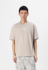 Beige T-Shirt mit kurzen Ärmeln und Rundhalsausschnitt, mit weißem "Hollister"-Schriftzug. Getragen mit hellgrauen gemusterten Shorts. Einfaches, lässiges Design.