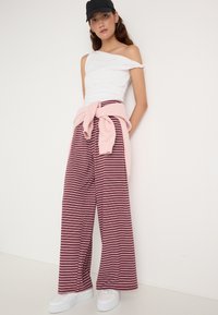 Mladá žena na sobě má bílé off-shoulder top, vínové pruhované široké kalhoty, růžový svetr uvázaný v pase, bílé tenisky a černou čepici.
