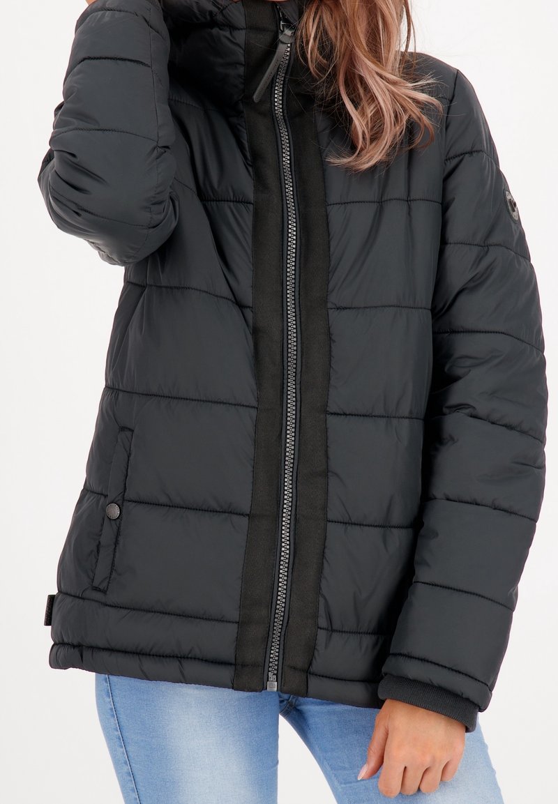 Vrouw in een zwarte pufferjack met zichtbare rits en zakken, gecombineerd met lichtblauwe jeans, staand tegen een effen achtergrond.