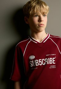 Burgundipunane T-särk valgete aktsentidega, millel on "SUB5CRIBE" logo ja geograafilised koordinaadid. Pehme kangas ning sportlik V-kaelus.