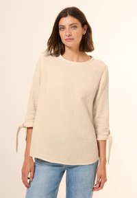 Beige Leinenoberteil mit Drei-Viertel-Ärmeln, rundem Ausschnitt und gebundenen Manschetten. Weiche Textur mit lockerer Passform. Getragen mit hellblauen Jeans.