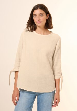 Beige Leinenoberteil mit Drei-Viertel-Ärmeln, rundem Ausschnitt und gebundenen Manschetten. Weiche Textur mit lockerer Passform. Getragen mit hellblauen Jeans.