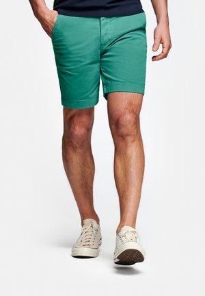 Man draagt groene korte broek en witte sneakers, loopt met één hand in de zak tegen een witte achtergrond.